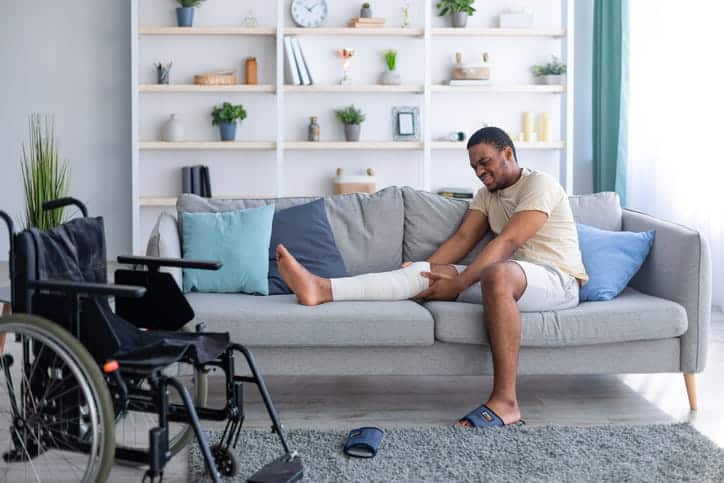 A man in pain on the couch with his leg in a cast. Next to his couch is a wheelchair. 