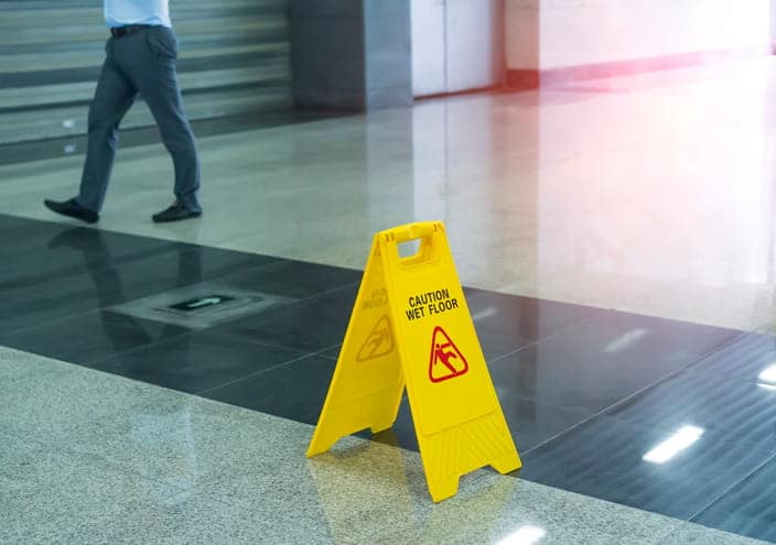 A yellow caution wet floor sign in a public space. In the background, a person is walking by.