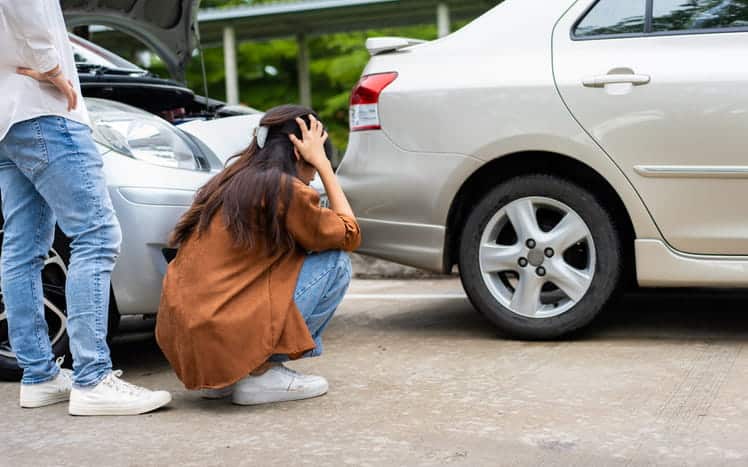 A woman squatting and holding her head in distress as she inspects the damage to her vehicle after a car accident.